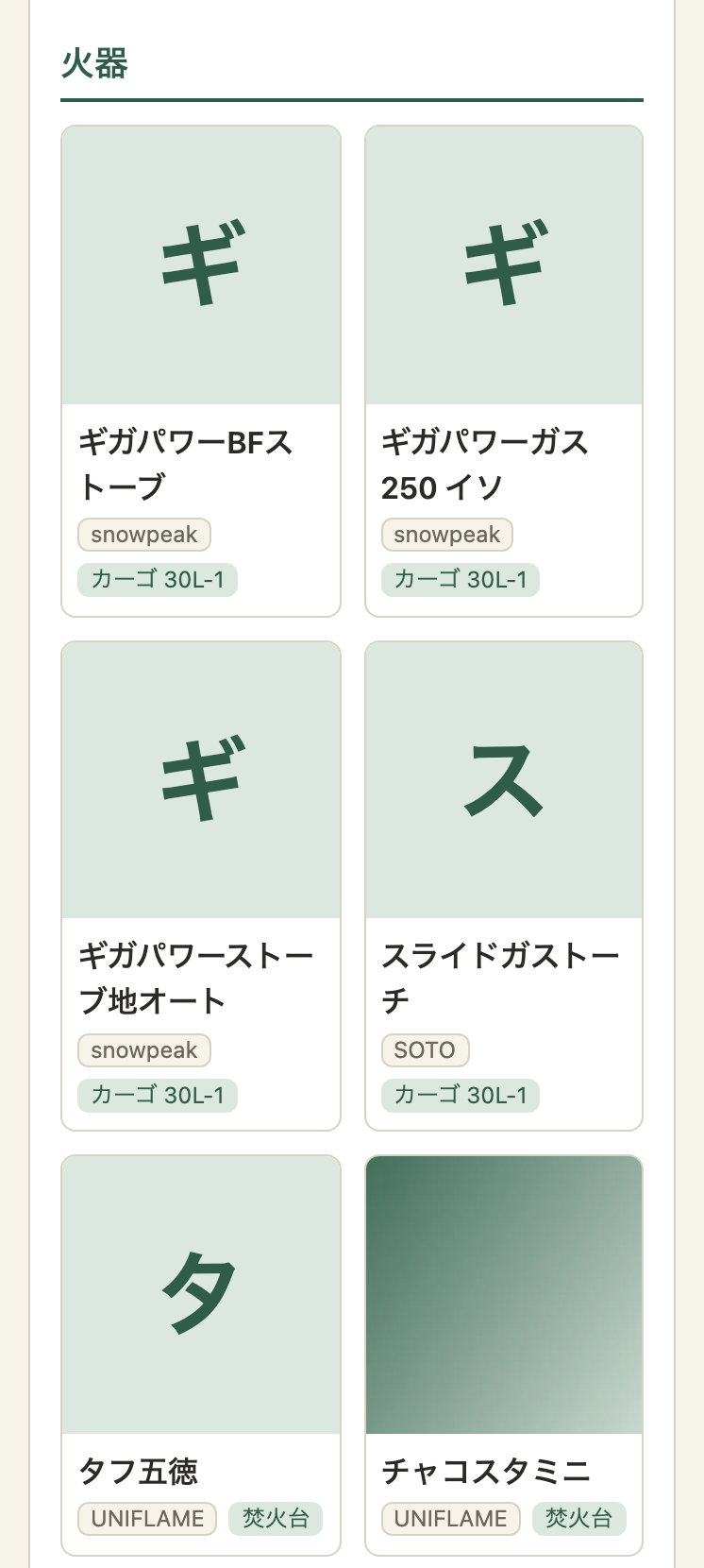 スマホの幅のギア一覧のカード表示。「火器」の見出しの下に、カードが2列で並ぶ。頭文字のカードの中に、緑のグラデーションの画像が付いたカードが1つある。長い製品名は2行に折れている